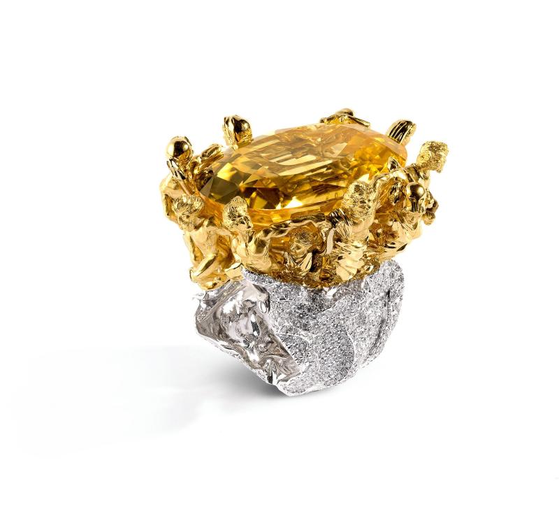 خاتم من الذهب الأبيض الخُلقي المرصّع بالألماس، يتوّج مركزه حجر ياقوتي أصفر زنة 127.70 قيراط، تُسنده منحوتات ذهبية عتيقة الطراز. 