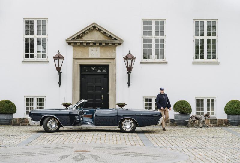 أندِرْس كيرك يوهانسن بجانب سيارته المكشوفة من طراز لينكولن كونتيننتال 1967 Lincoln Continental ومعه كلبه بيبي، من فصيلة "أولْد دانيش بويْنتر"، خارج المنزل الرئيس في إقامة رودن غادْس. 